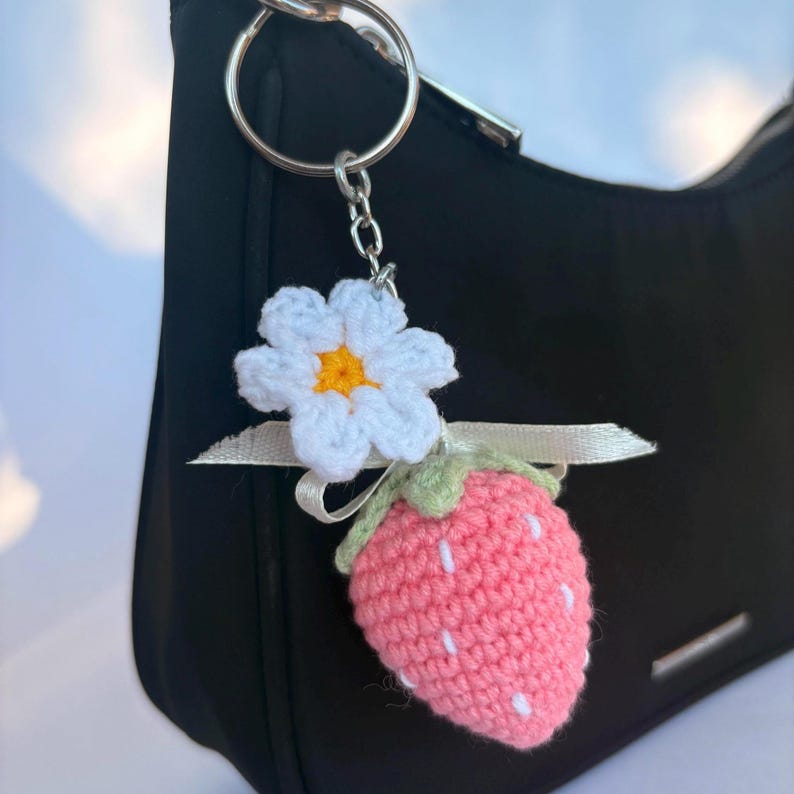 May include: A crochet keychain featuring a pink strawberry with white embroidered seeds, a green stem, and a white flower with a yellow center. A thin white ribbon is tied between the flower and the strawberry, all attached to a silver metal ring.