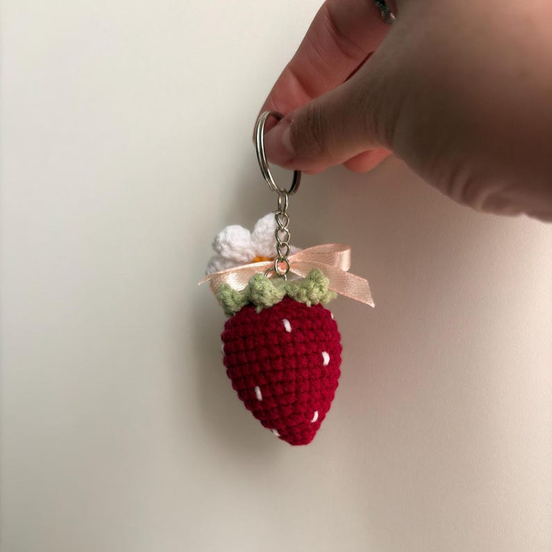 May include: A crochet strawberry keychain featuring a red berry with white embroidered seeds, a green calyx, a small white flower with a yellow center, and a light pink ribbon bow attached to a silver metal split ring for keys.