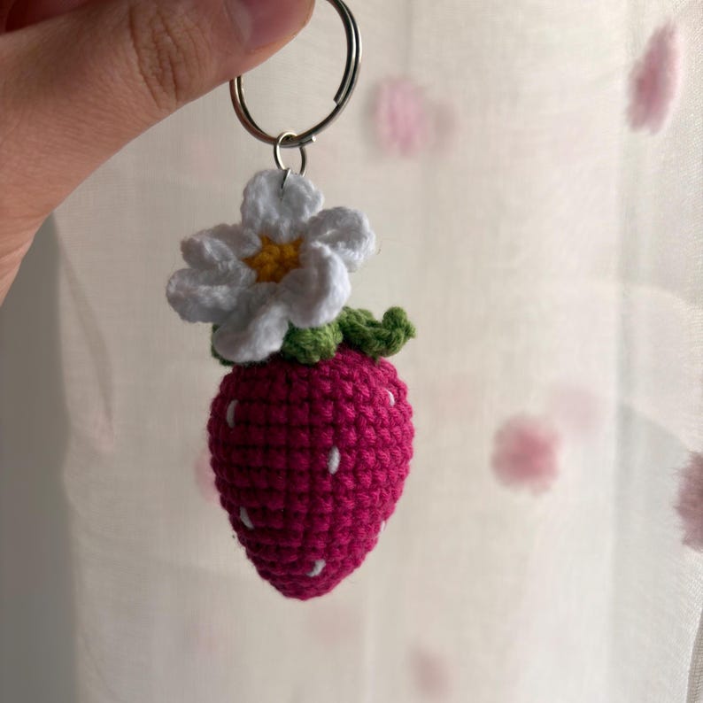 May include: A crocheted strawberry keychain featuring a bright pink berry body with small white embroidered accents, topped with a green crocheted stem and a white flower with a yellow center, all attached to a silver metal split ring.