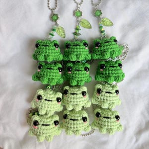 May include: Three vertical hanging chains featuring crocheted green frogs. Each chain has four frogs in varying shades of green, from bright to pale, with black bead eyes and pink cheeks. The chains are accented with small green leaf charms and beads.