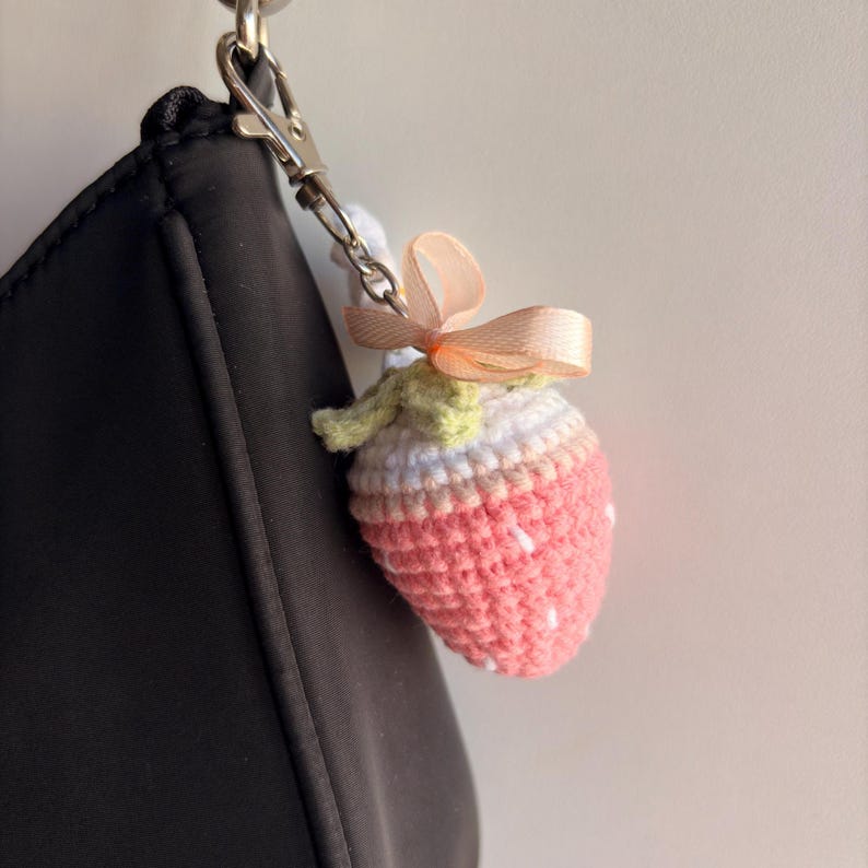 May include: A crochet strawberry keychain featuring a pink body with white embroidered seeds, a white top section, and a light green stem. The strawberry is adorned with a small peach-colored ribbon bow and attached to a silver metal lobster clasp.