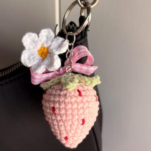 May include: A crochet strawberry keychain featuring a light pink textured body with small red embroidered seeds, a green stem, a white flower with a yellow center, and a pink and white gingham ribbon bow attached to a silver metal keyring.