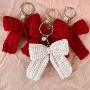 Pode incluir: Três porta-chaves de laço em croché, dois em vermelho escuro e um em branco. Cada laço é feito com um ponto de fio texturizado e possui um anel de metal prateado preso a uma corrente curta com uma pequena conta redonda de cor creme.