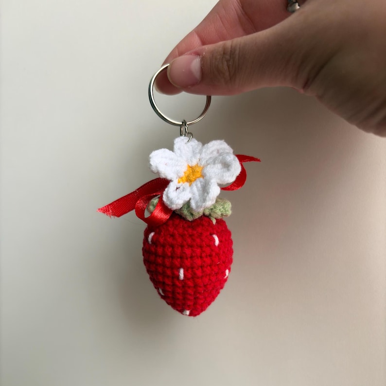 May include: A crocheted strawberry keychain featuring a bright red textured body with small white stitched details, topped with a white and yellow crocheted flower, a small green leaf accent, and a thin red ribbon tied to a silver metal ring.
