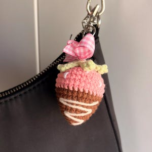 May include: A crochet strawberry charm featuring a pink top, a brown chocolate-dipped bottom with white yarn drizzle, and a light green stem. A small pink and white gingham ribbon bow is attached to the silver metal lobster clasp hardware.
