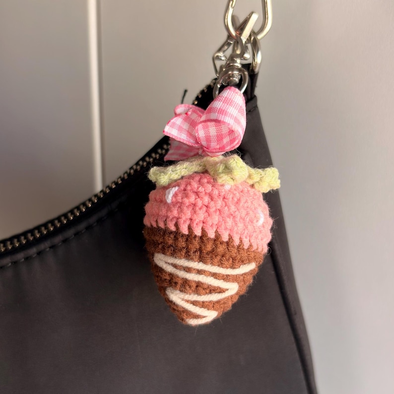 May include: A crochet strawberry charm featuring a pink top, a brown chocolate-dipped bottom with white yarn drizzle, and a light green stem. A small pink and white gingham ribbon bow is attached to the silver metal lobster clasp hardware.