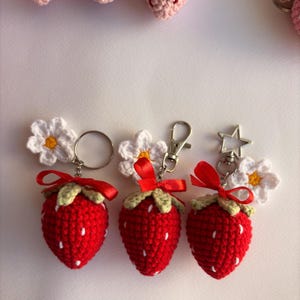 May include: Three crocheted strawberry keychains, each featuring a red berry with white embroidered seeds, a light green top, and a small red ribbon bow. Each strawberry is attached to a white crocheted flower and a metal ring or clasp.