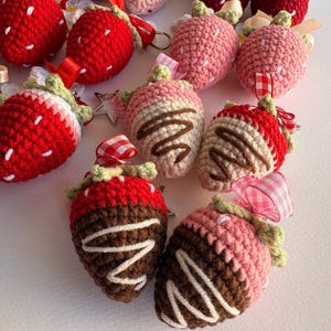May include: A collection of crocheted strawberry keychains in various colors including bright red, light pink, and chocolate-dipped designs. Each strawberry features a green crocheted stem, a small ribbon loop, and a metal keyring attachment.