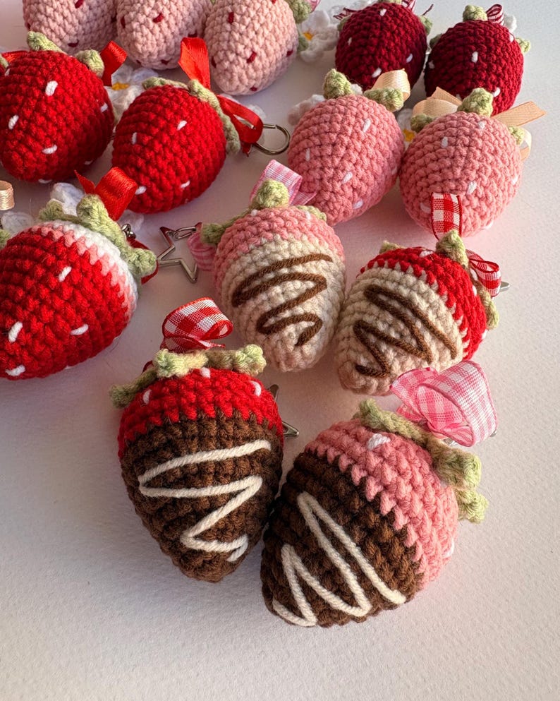 May include: A collection of crocheted strawberry keychains in various colors including bright red, light pink, and chocolate-dipped designs. Each strawberry features a green crocheted stem, a small ribbon loop, and a metal keyring attachment.
