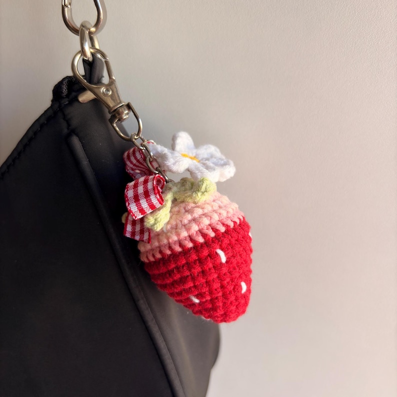 May include: A crochet strawberry keychain featuring a red body with white embroidered seeds, a light pink top, and green leaves. It is adorned with a small white flower and a red and white checkered ribbon, attached to a silver metal lobster clasp.