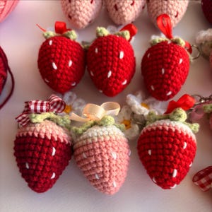 May include: A collection of crocheted strawberry keychains featuring red and light pink yarn bodies with white embroidered seeds, green yarn leaves, and small fabric ribbons in red, white gingham, and beige attached to metal key rings.