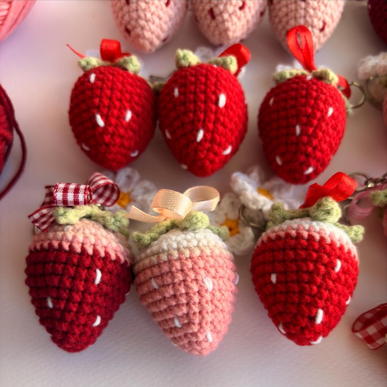 May include: A collection of crocheted strawberry keychains featuring red and light pink yarn bodies with white embroidered seeds, green yarn leaves, and small fabric ribbons in red, white gingham, and beige attached to metal key rings.