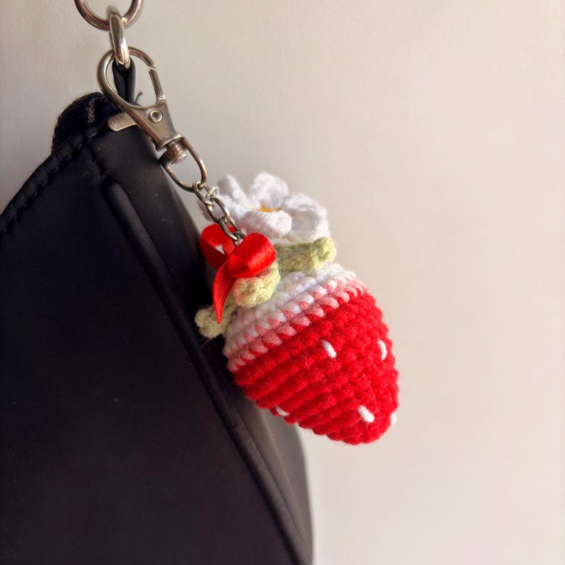 May include: A crochet strawberry keychain featuring a red fruit body with white embroidered seeds, a green stem, and a white flower topper. A small red ribbon bow is tied near the silver-toned metal lobster clasp attachment.