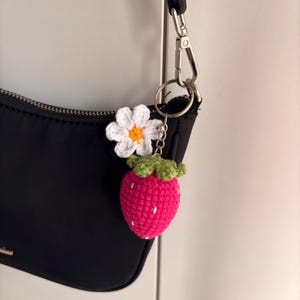 May include: A crochet strawberry keychain with a white daisy flower attached to a silver metal ring. The strawberry is bright pink with small white embroidered seeds and a green crocheted top, hanging from a black fabric shoulder bag.