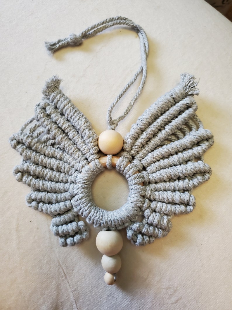 K&ouml;nnte beinhalten: Ein grauer Makramee-Engel-Fl&uuml;gel-Wandbehang mit einem Holzring und drei Holzperlen.