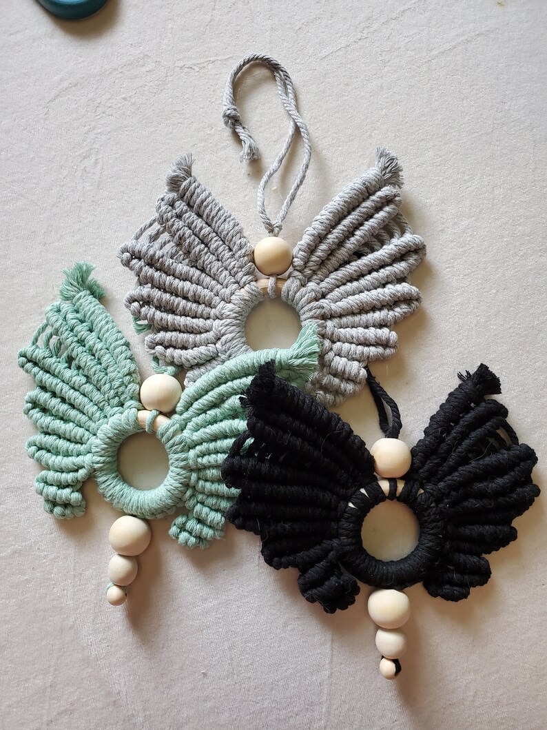 K&ouml;nnte beinhalten: Drei Makramee-Schmetterlings-Wandbeh&auml;nge mit Holzperlen. Die Schmetterlinge sind in Grau, Gr&uuml;n und Schwarz.