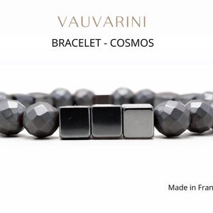 Puede incluir: Una pulsera gris oscuro con cuentas esféricas facetadas y cuentas cuadradas. La pulsera está etiquetada como "BRACELET - COSMOS" y "Made in France". Las cuentas son de un color gris oscuro uniforme.