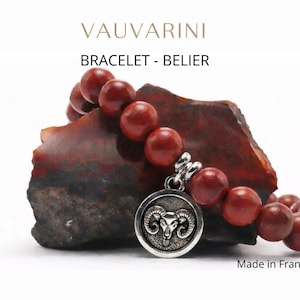 Peut inclure: Un bracelet de perles rouges avec un charm en argent représentant une tête de bélier. Le bracelet est présenté sur une pierre sombre et texturée. Le texte "VAUVARINI" et "BRACELET - BELIER" est visible. "Made in France" est également présent.