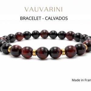 Puede incluir: Una pulsera de cuentas con cuentas rojas oscuras y negras alternas, acentuada con espaciadores dorados. La pulsera está etiquetada como "BRACELET - CALVADOS" y "Made in France".