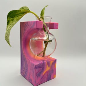 May include: A decorative plant propagation station with a clear glass orb and a small top cup, holding a plant cutting. The station is made of a block of pink, purple, and orange swirled resin. The plant has green and yellow variegated leaves.
