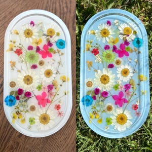May include: Two oval resin trays, one white and one light blue, each embedded with colorful dried flowers. The flowers include white daisies, pink, yellow, and blue blossoms. The trays are decorative and suitable for holding small items.