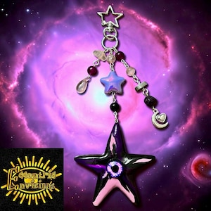 May include: A celestial-themed keychain with a large, dark purple star pendant featuring a light blue eye design. The keychain includes a silver star-shaped clasp, smaller star charms, and various beads in shades of pink, purple, and clear. The background is a nebula.