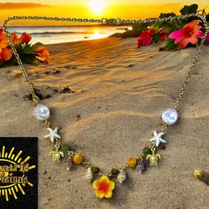 May include: A gold-colored necklace with a beach theme, featuring pearls, starfish, sea turtles, and a yellow and orange flower pendant. The necklace is displayed on sand with flowers in the background.