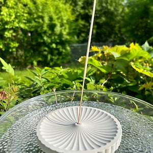 May include: A white ceramic incense holder with a fan-shaped design. The holder has a small hole in the centre for an incense stick. The incense stick is burning and producing smoke.