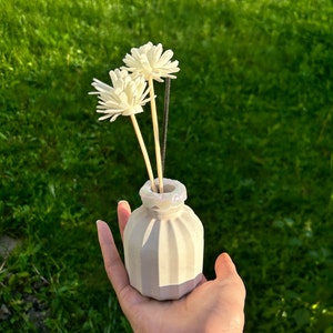 May include: A white ceramic diffuser bottle with a textured surface and an iridescent glitter rim. The bottle is filled with wooden sticks and topped with three white artificial flowers.