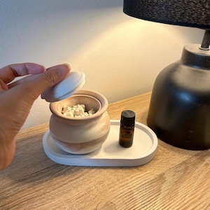 May include: A white ceramic diffuser with a lid, filled with white diffuser stones, and a small brown glass bottle of essential oil. The diffuser is sitting on a white oval tray.