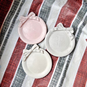 May include: Three decorative ceramic dishes with a bow design. The dishes are in shades of pink, grey, and white.
