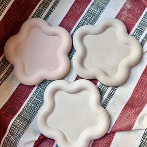May include: Three ceramic flower-shaped dishes in shades of pink and beige. The dishes are arranged on a striped fabric background.