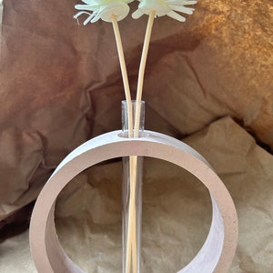 May include: A white ceramic vase with a circular opening, holding a glass tube with two white flower stems and wooden sticks.