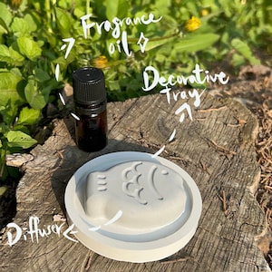 May include: A white ceramic diffuser shaped like a fish with a small brown glass bottle of fragrance oil on a wooden surface.