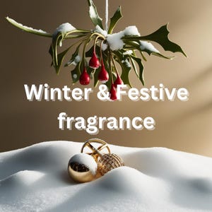 May include: A sprig of holly with red berries is covered in snow and hangs over two gold Christmas ornaments on a white snowy surface. The text "Winter & Festive fragrance" is written below the holly.