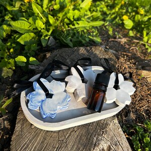 May include: Three white ceramic flower-shaped air fresheners with blue accents, a small brown glass bottle of essential oil, and a grey concrete tray. The items are on a tree stump.