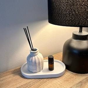 May include: A white ceramic diffuser with black reeds, a small brown glass bottle of essential oil, and a white ceramic tray. The diffuser and bottle are on the tray. The tray is on a wooden surface.