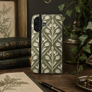 May include: A sage green phone case featuring a repeating cream-colored botanical pattern of Venus flytraps. The case has a vertical black elastic strap running down the left side, covering the camera lens area, and a textured, matte finish.