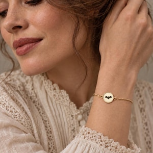 May include: A gold-toned chain bracelet featuring a circular charm with a black bat silhouette engraved in the center. The delicate metal chain connects to the round pendant, which displays a detailed dark bat graphic against a light background.