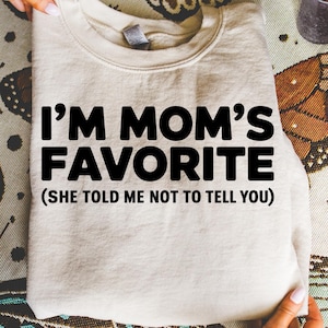 May include: A beige sweatshirt with the text "I'M MOM'S FAVORITE (SHE TOLD ME NOT TO TELL YOU)" in bold black letters. The folded sweatshirt is on a patterned surface.