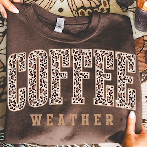 May include: A brown sweatshirt with the words "COFFEE WEATHER" in a bold, leopard print design. The sweatshirt is folded, and the text is centered on the front. The overall theme is coffee and cozy weather.