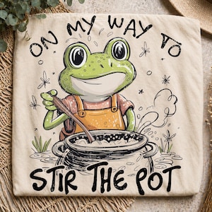May include: Beige tote bag featuring a cartoon frog wearing an orange apron and stirring a pot. The text "ON MY WAY TO STIR THE POT" is printed in black. The frog is green with a smiling expression.