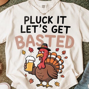 Puede incluir: Camiseta color crema con el texto "PLUCK IT LET'S GET BASTED". Un pavo de dibujos animados con sombrero y sujetando una jarra de cerveza aparece debajo del texto, rodeado de hojas de otoño. La camiseta es de la marca Comfort Colors.