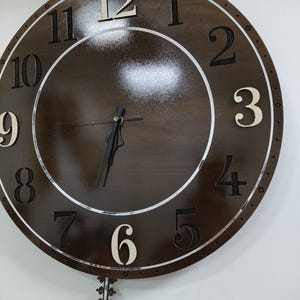 May include: A large, round wooden wall clock with a dark brown face and white numerals. The clock has a silver-colored ring around the face and a decorative pendulum. The hands are black.