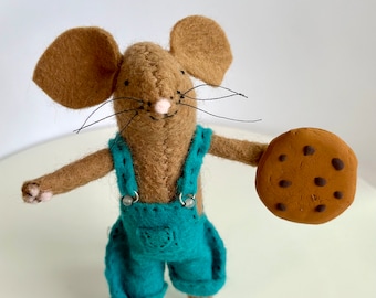 May include: A brown felt mouse wearing blue overalls holds a chocolate chip cookie in its right hand. The mouse has a pink nose and black whiskers.