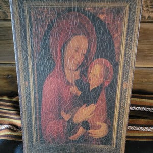 May include: A vintage-style framed painting of a mother and child. The painting is done in a warm palette of browns, reds, and yellows. The mother is wearing a red robe and the child is wearing a dark robe. The painting has a cracked and aged look, giving it a rustic feel.