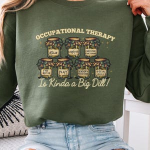 May include: Olive green sweatshirt with the text "Occupational Therapy Is Kinda a Big Dill!" and illustrations of jars labeled with OT concepts. The sweatshirt is a casual, comfortable style.