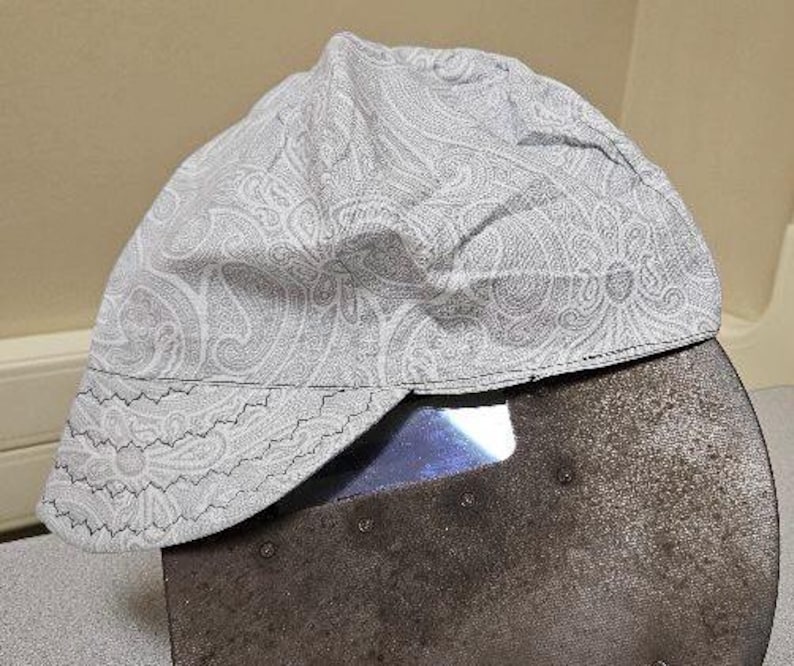 May include: A white welding cap with a paisley pattern. The cap has a brim and a back flap that can be folded down.