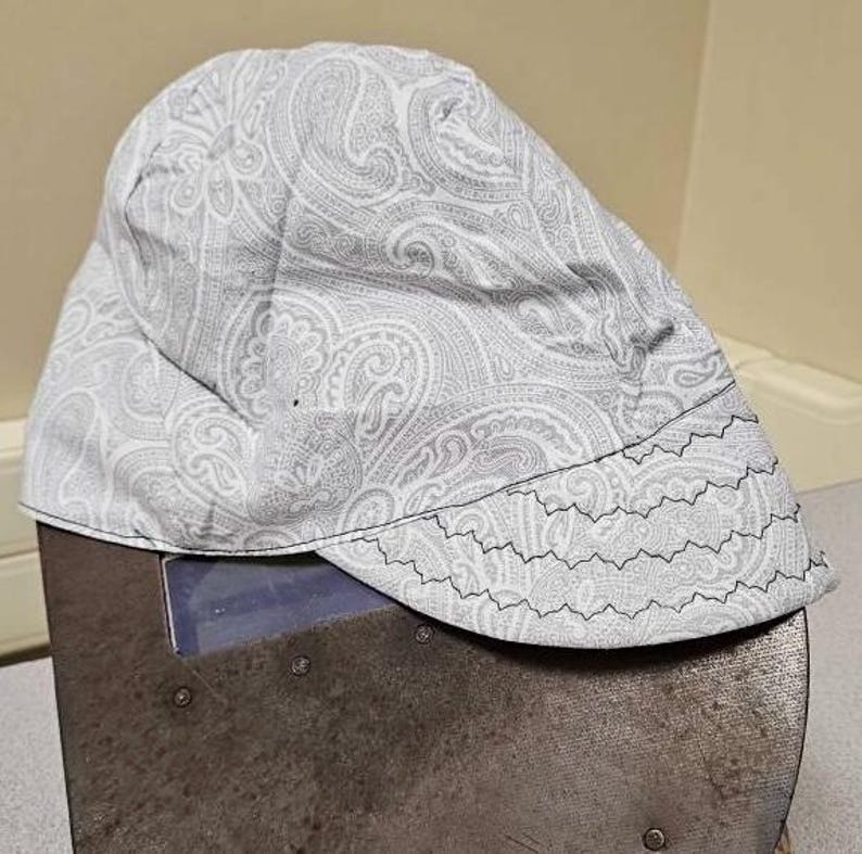 May include: A white paisley patterned welding cap with a black zig-zag stitch around the brim.