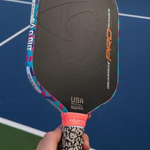 May include: A black pickleball paddle with a matte surface, featuring the text SST CORE, GEARBOX PRO POWER, and USA PICKLEBALL APPROVED. The edge guard has a vibrant blue and pink splatter pattern, and the handle has a grey and black elephant print.
